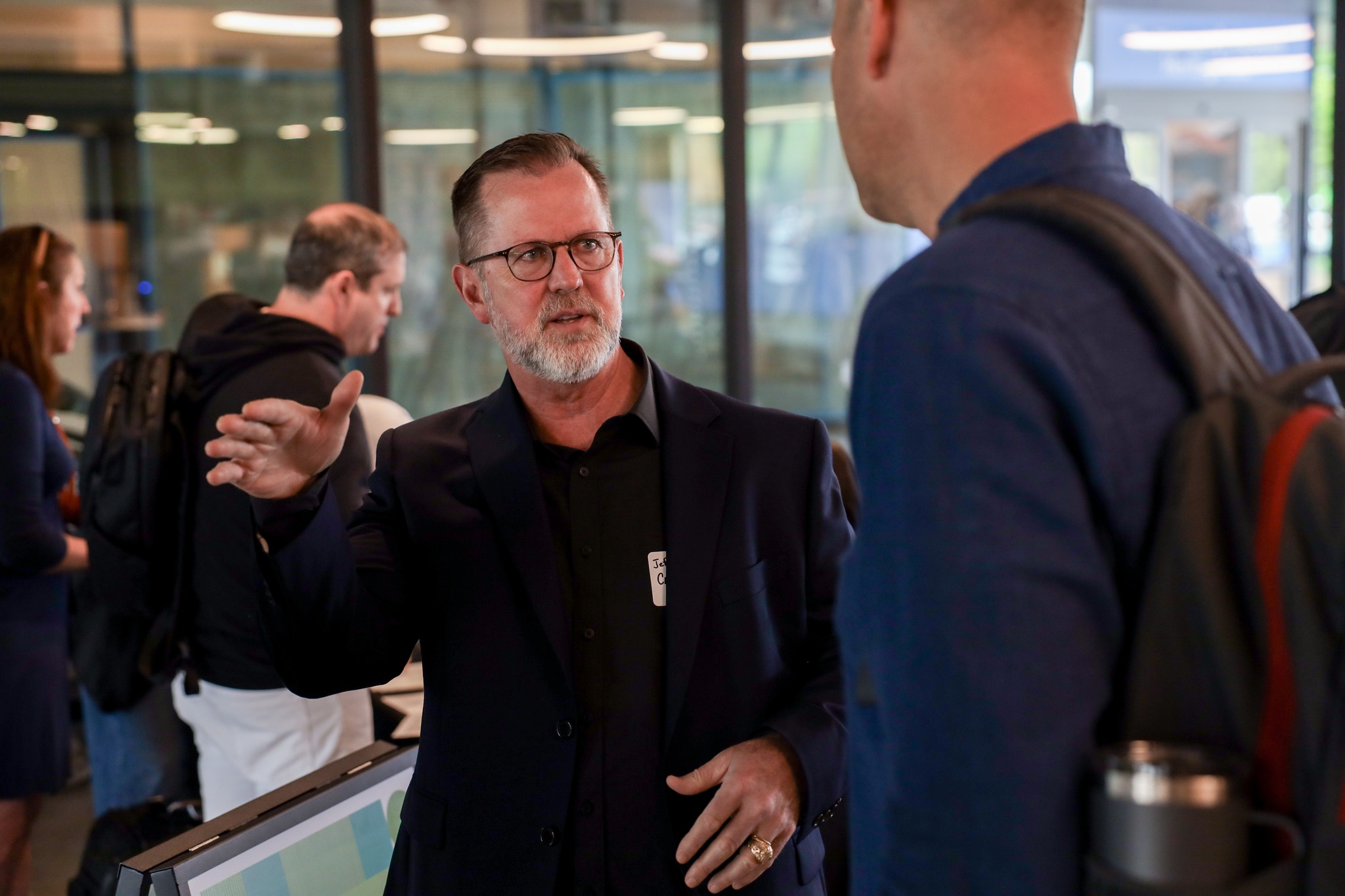 그물 A man in a dark suit and glasses gestures while speaking to another man in a blue shirt inside a modern, glass-walled office space. Other people are visible in the background.