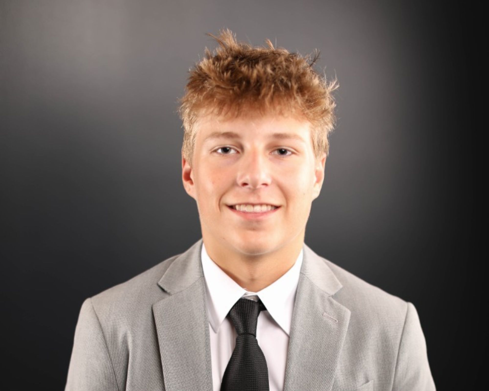 A young person with short, tousled blonde hair, wearing a light gray suit, white shirt, and black tie, is smiling in front of a plain dark background.