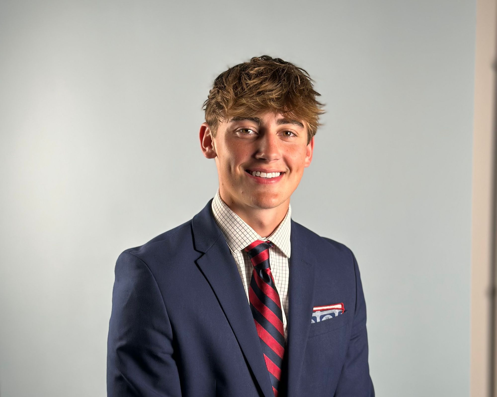 A young man with light brown hair wearing a navy blue suit, checkered shirt, and a red striped tie, smiling in front of a plain light gray background.
