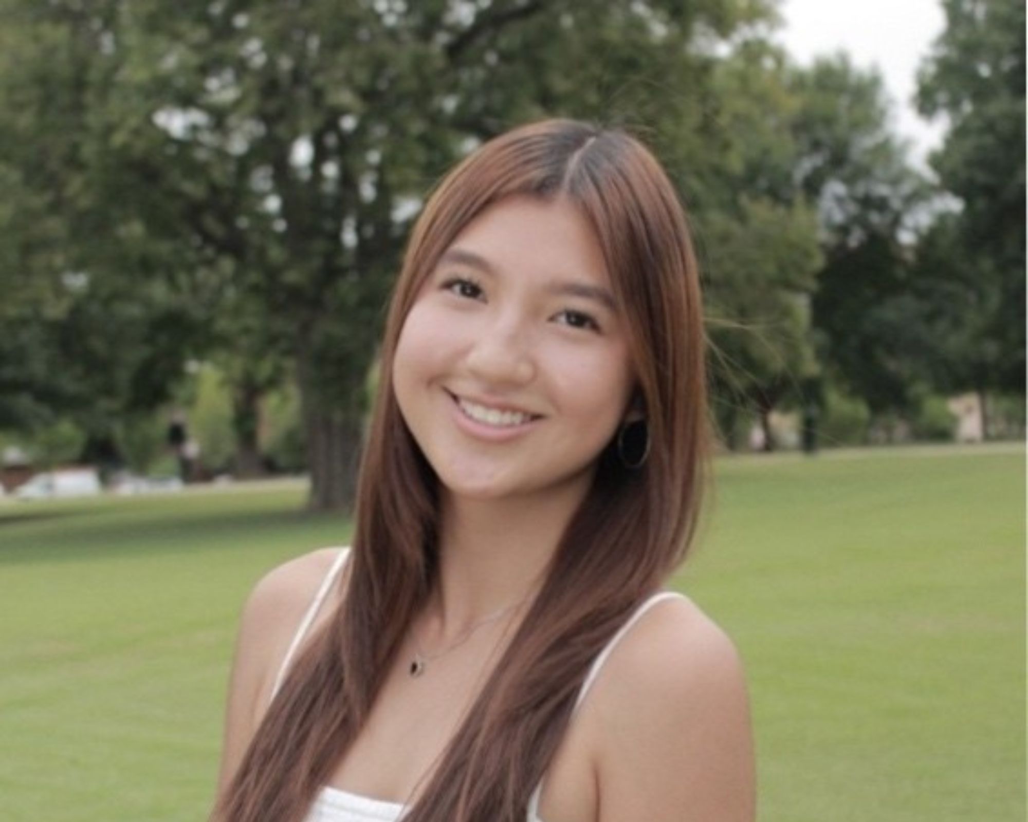 A young woman with long brown hair smiles at the camera while standing outdoors on a grassy field, with trees and a cloudy sky in the background. She wears a white sleeveless top and a necklace.