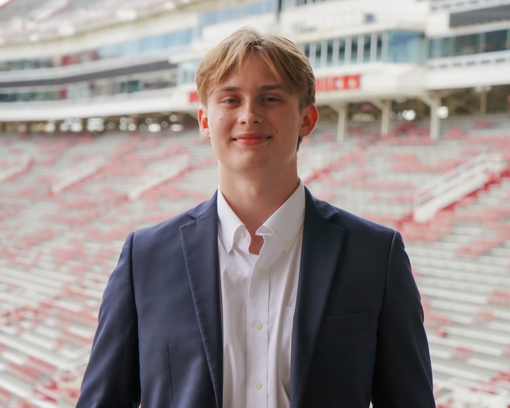 A young man in a navy suit and white dress shirt stands smiling in front of empty stadium seats arranged in rows. The background is blurred, focusing on the man.