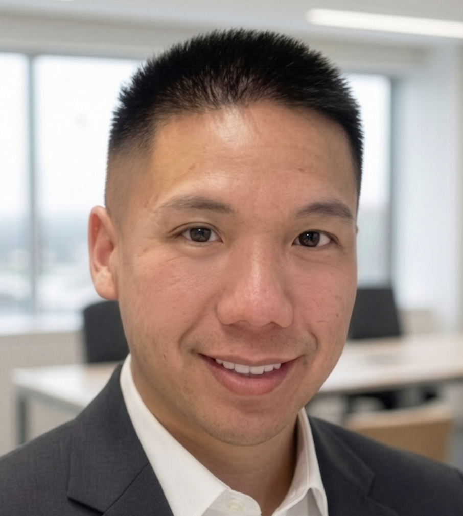A man with short black hair, wearing a dark suit jacket and white shirt, smiles at the camera in a modern office with large windows and blurred desks in the background.