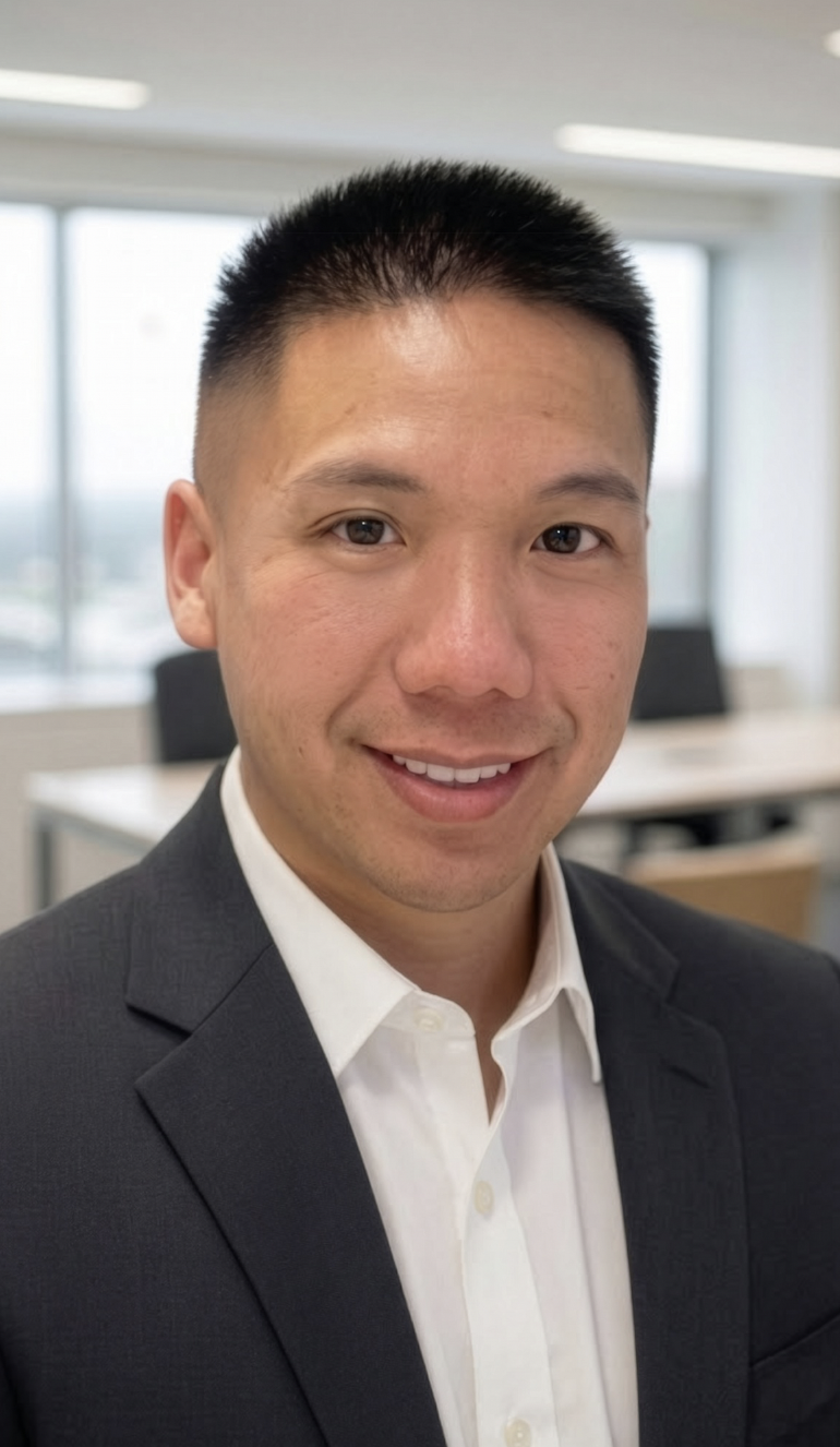 A man wearing a dark suit and white dress shirt smiles at the camera in a modern office with large windows and desks in the background.
