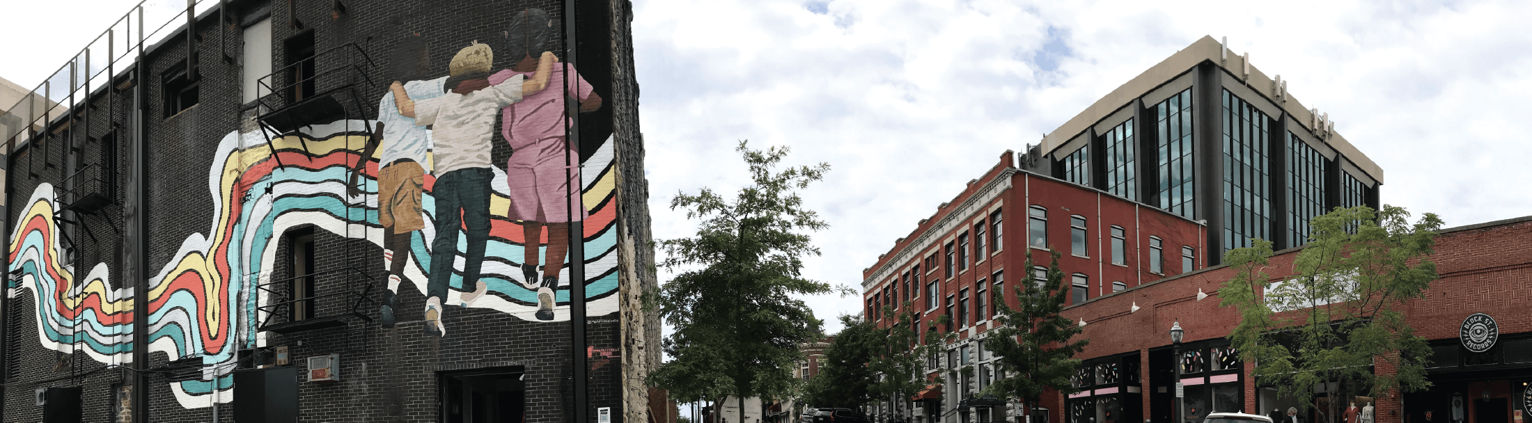 A mural of three people holding hands and jumping, with colorful wavy lines behind them, decorates the side of a dark brick building; nearby are red brick buildings and trees under a cloudy sky.
