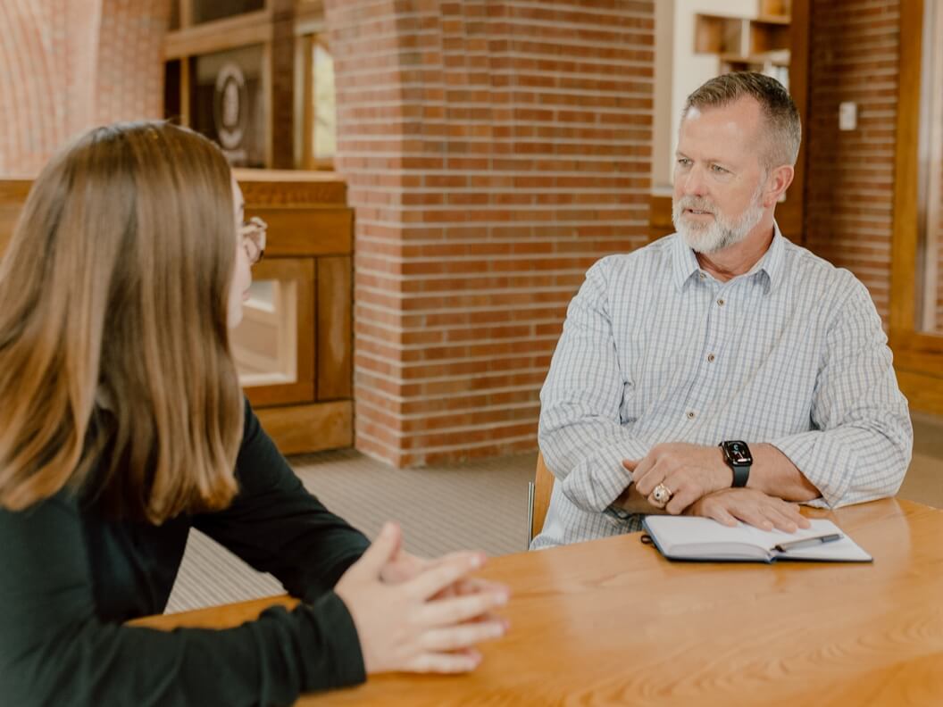 A man and a woman sit across from each other at a wooden table, engaged in conversation. The man has a notepad open in front of him, and both are inside a room with brick walls and large windows.