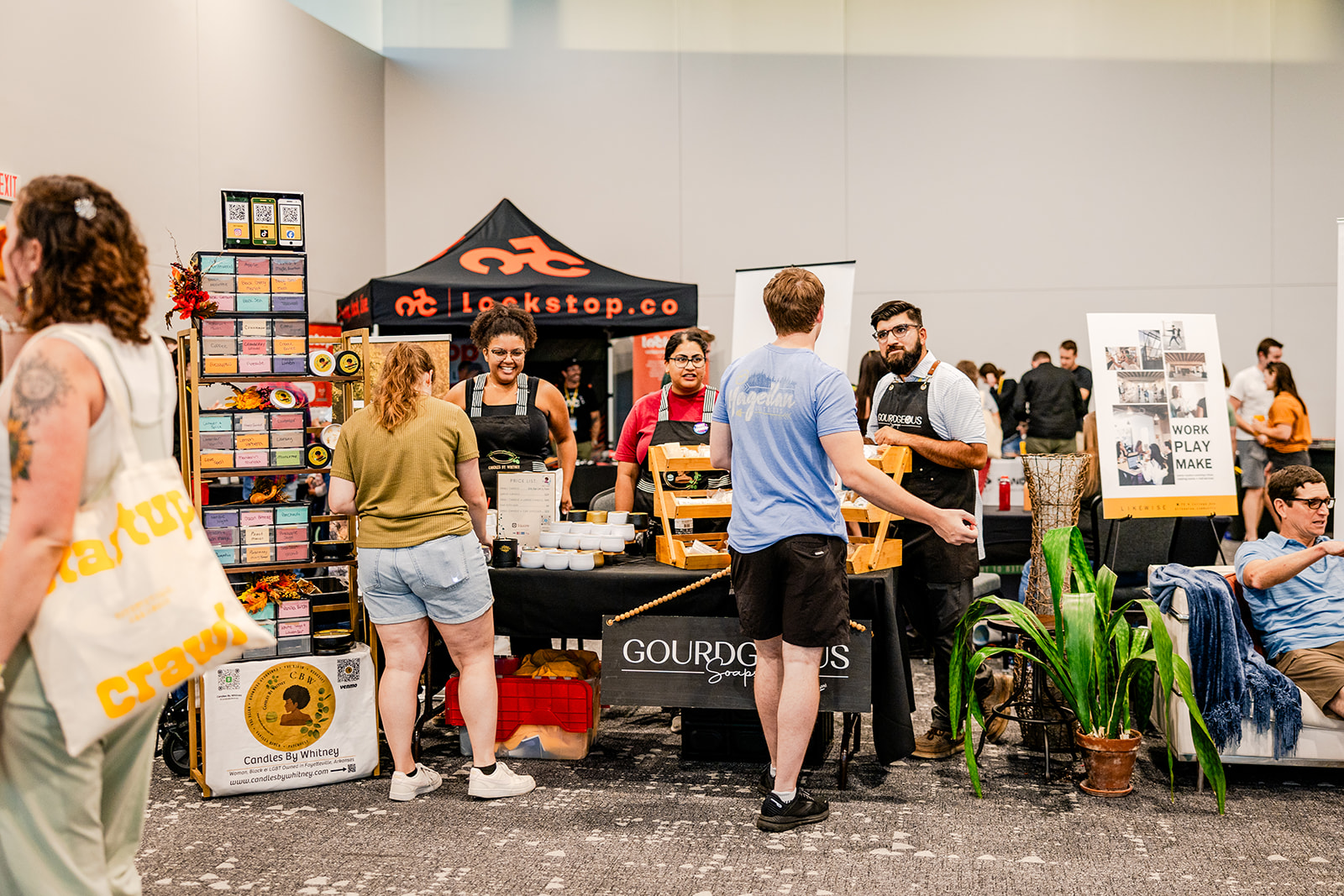 People interact at a bustling indoor market with colorful booths, a display of goods, and informational signs. A black canopy tent and several vendors stand behind tables with products. Shoppers browse and chat in the scene.