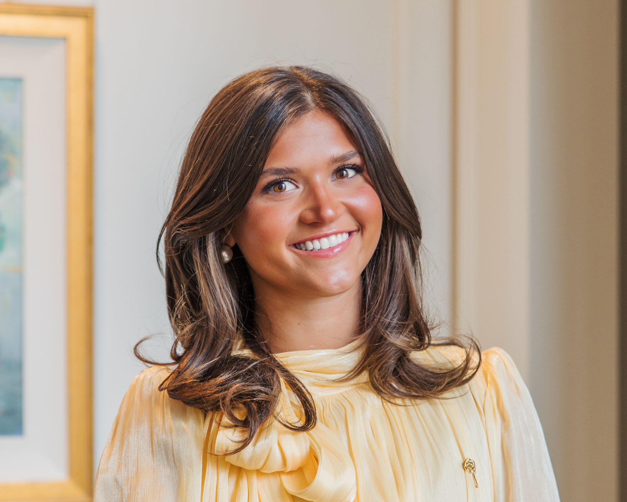 A young woman with medium-length brown hair, wearing a pale yellow blouse with a bow, smiles at the camera. She stands indoors near a white wall and a framed picture.