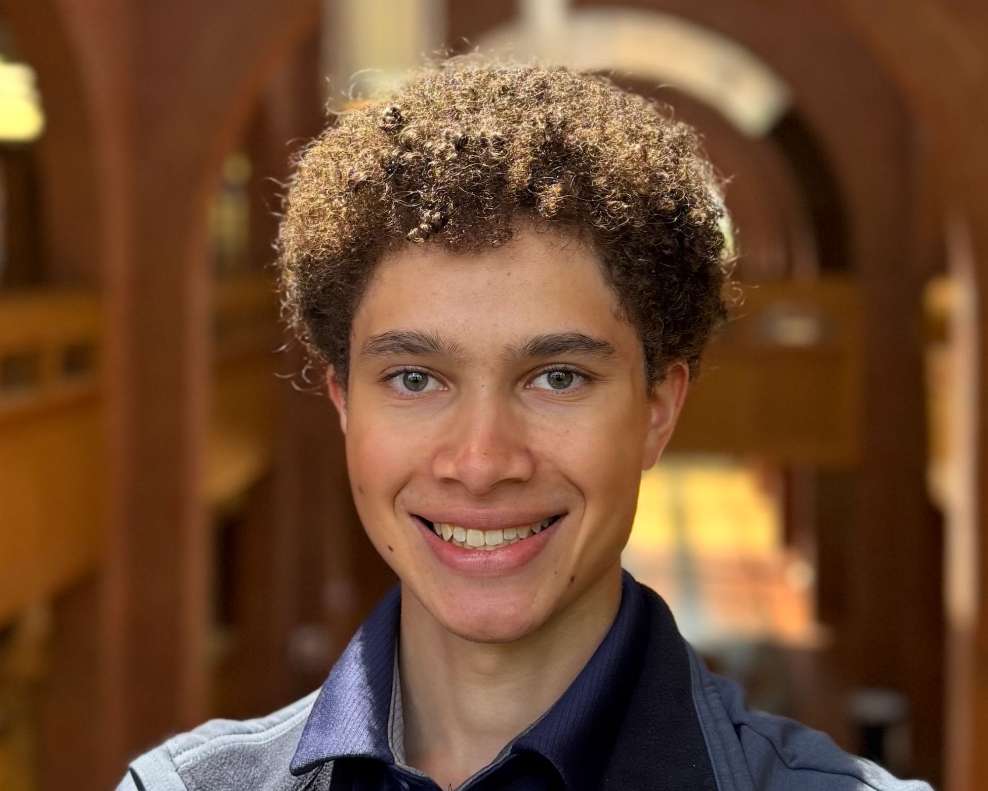 A young person with curly hair smiles at the camera, standing indoors in front of blurred, arched architectural details.