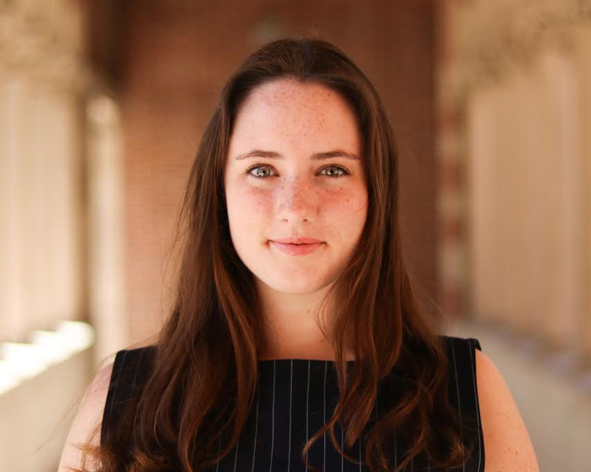 A young woman with long brown hair and freckles stands in a sunlit corridor, wearing a navy blue top with subtle pinstripes, looking directly at the camera with a soft, neutral expression.