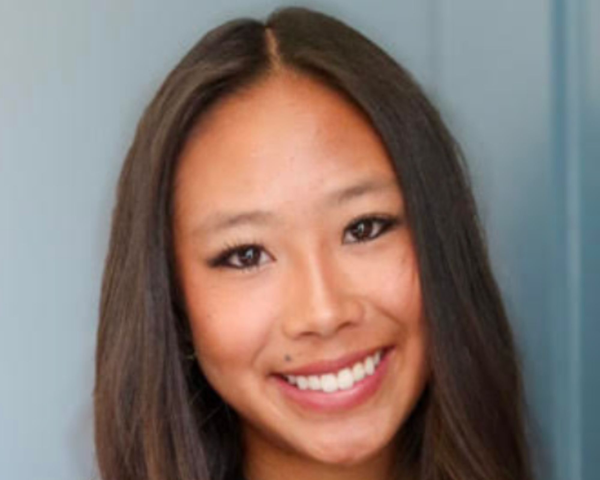 A young woman with long, straight brown hair smiles at the camera in front of a blue background.