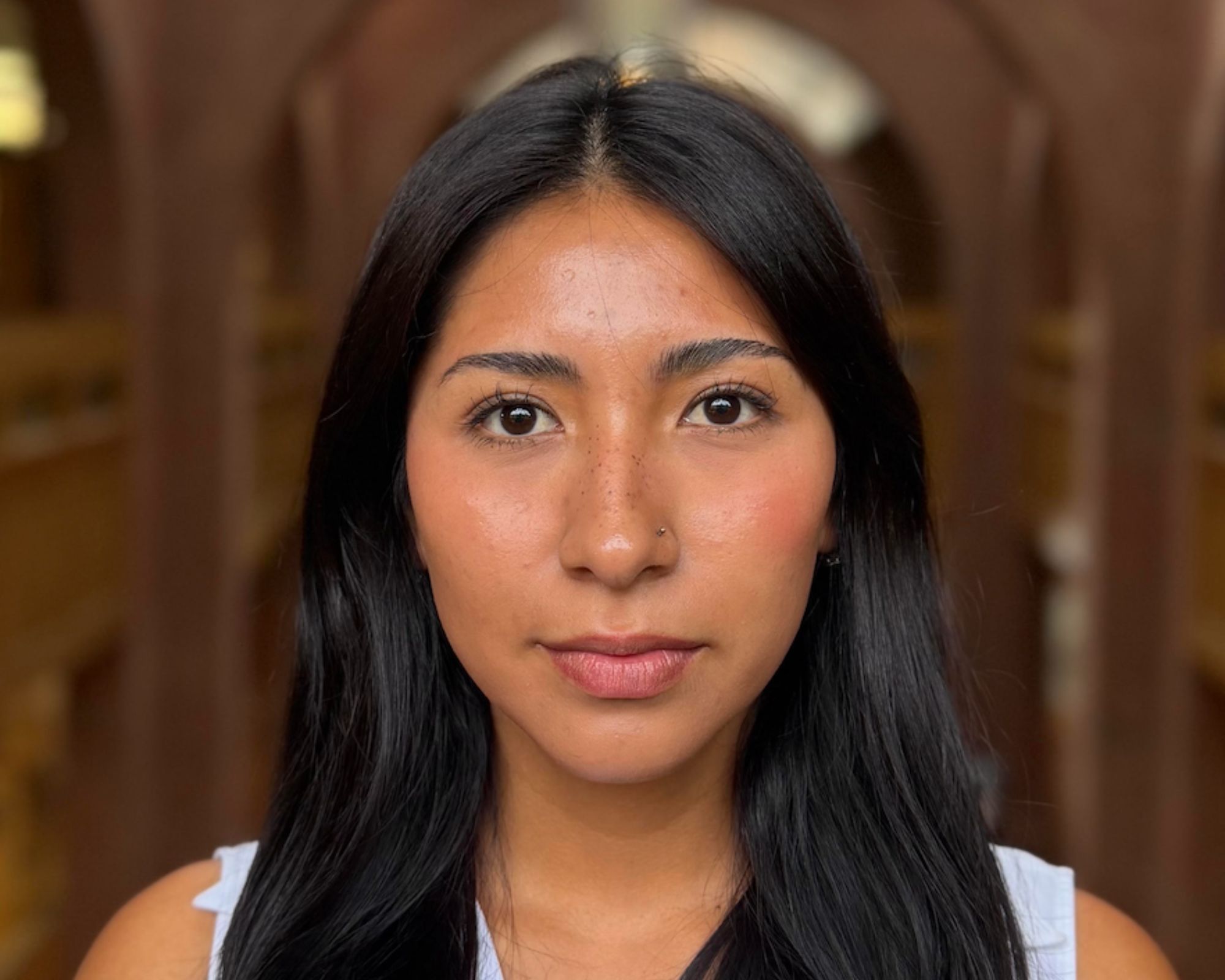A woman with long black hair and a nose piercing looks directly at the camera, standing in a softly lit indoor space with blurred architectural details in the background.