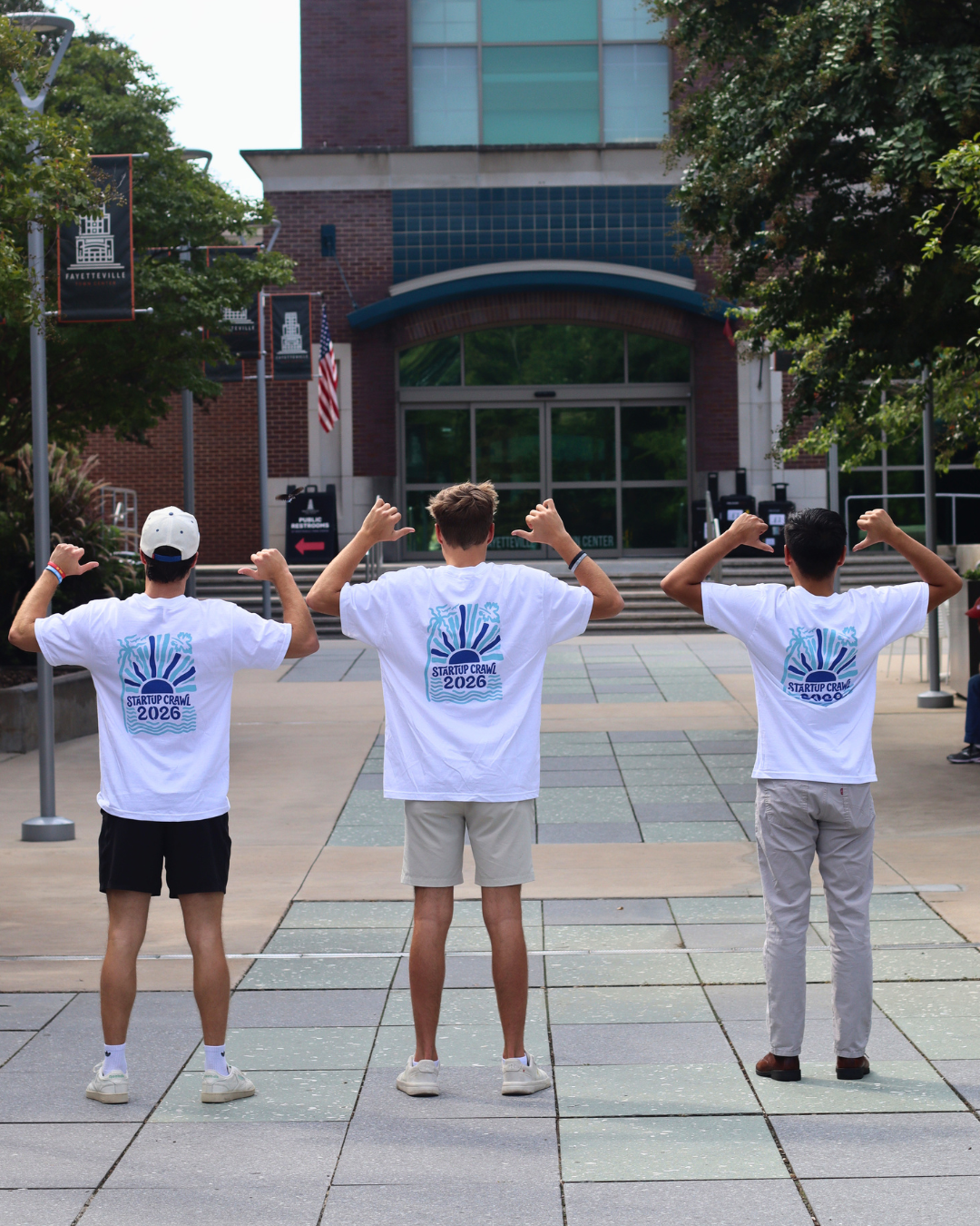 Three people stand outside, facing away, wearing matching white T-shirts that say Explore 2026 in blue and green. They point at the designs on their backs on a paved walkway in front of a building.