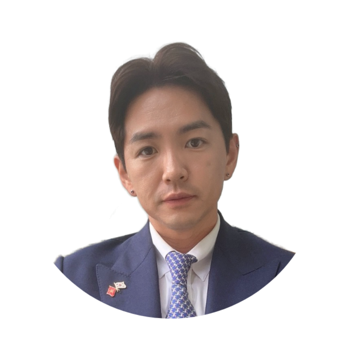 A person with short dark hair in a blue suit and patterned tie, wearing earrings, is looking at the camera against a white background.