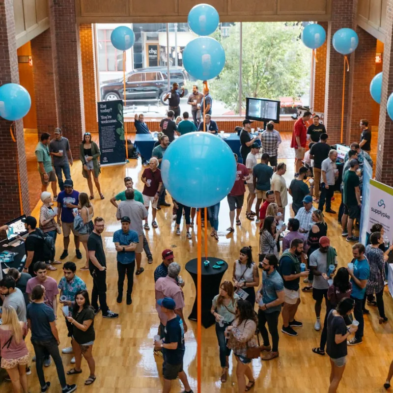 A large group of people socializes and explores booths at an indoor Startup Crawl event with blue balloons and tall windows.