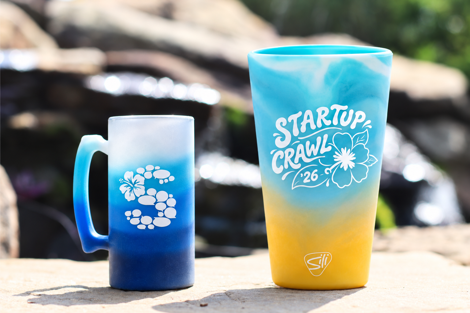 Two colorful gradient cups with tropical designs sit on a sunlit rock outdoors; one says Startup Crawl 26 with a hibiscus, the other has a floral pattern. A blurred waterfall and greenery are in the background.