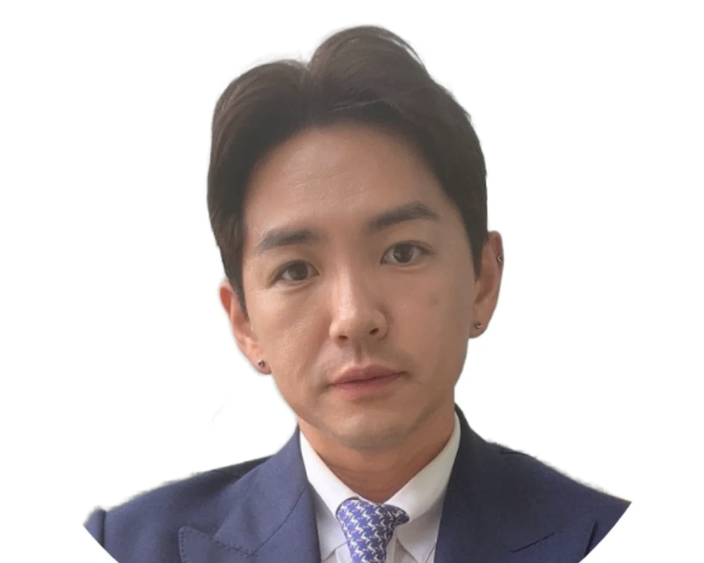 A person with short dark hair in a blue suit and patterned tie, wearing earrings, is looking at the camera against a white background.